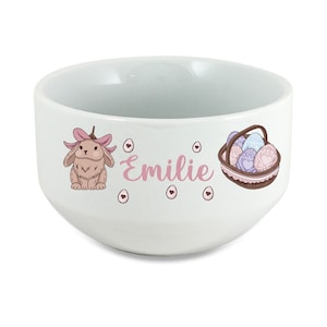 Peut inclure: Bol en céramique blanche avec un lapin rose, des œufs roses et un panier d'œufs. Le bol porte le nom "Emilie".