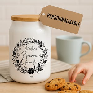 Peut inclure: Pot en céramique blanc avec couvercle en bois, orné d'une couronne florale noire et de texte. Une étiquette indique "PERSONNALISABLE". Des cookies aux pépites de chocolat et une tasse bleue sont à proximité. Le pot est un article de rangement de cuisine.