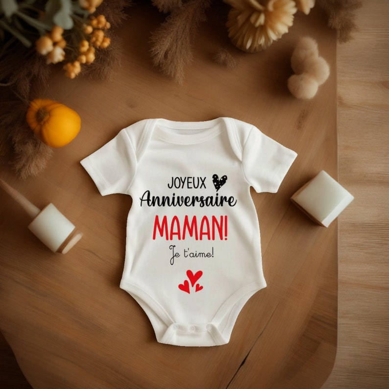 May include: White baby onesie with black text that reads "Joyeux Anniversaire Maman! Je t'aime!" with a black and white heart and three red hearts.