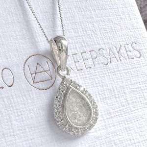May include: A silver teardrop pendant necklace featuring a central textured white inlay surrounded by a halo of small clear stones. The pendant hangs from a delicate silver chain against a white textured background with the text KEEPSAKES.