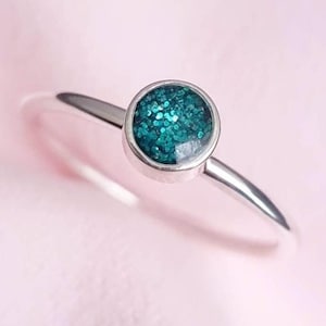 May include: A silver ring featuring a round teal glitter stone. The ring has a slender, polished band, and the stone is set in a simple bezel. The background is a soft pink.