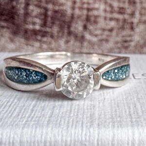 May include: A silver ring with a large, clear, round gemstone at the centre. The band features blue-green inlays on either side of the central stone. The ring is set on a white surface.