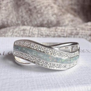 May include: A silver-toned ring featuring a central wavy inlay of iridescent, light blue crushed material flanked by two rows of small, clear, sparkling stones. The band has a polished metallic finish and a crossover design that curves across the centre.