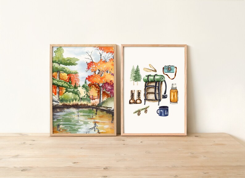 Op de afbeelding: Twee ingelijste aquarellen. De eerste aquarel toont een sc&egrave;ne met een meer en bomen in herfstkleuren. De tweede aquarel toont een verzameling kampeeruitrusting, waaronder een rugzak, laarzen, een thermoskan, een mok en een camera.