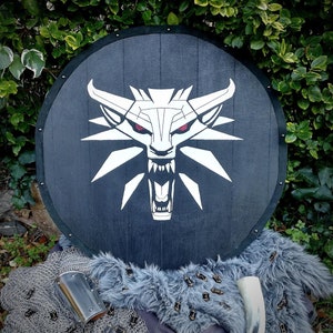 Op de afbeelding: Een zwart houten schild met een wit wolvenkopembleem. De wolf heeft rode ogen en een grommende uitdrukking. Het schild is omgeven door een kettinghemd, een pels en een hoorn.