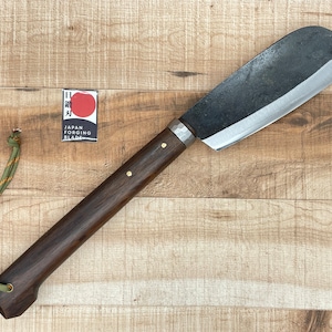 May include: A long, curved, black blade with a wooden handle and a brown leather strap. The blade is made in Japan and has a label that says "Japan Forging Blade".