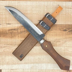 Può includere: Un coltello artigianale con un manico in legno scuro e una lama argentata, appoggiato su un fodero in legno. Il fodero è fissato con due cinghie in pelle nera. Il coltello è progettato per tagliare e tritare.