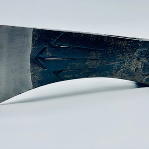 May include: Close-up of a metal axe head with a sharp, silver blade. The dark, textured body features an arrow design and a stamped emblem. The axe head is designed for chopping wood and other outdoor tasks.