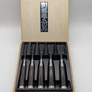 Peut inclure: Un ensemble de six ciseaux à bois avec des manches marron foncé et des accents argentés, disposés dans un plateau noir à l'intérieur d'une boîte en bois clair. Le couvercle de la boîte porte du texte japonais argenté. Pour le travail du bois.