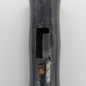 Può includere: Un utensile metallico cilindrico grigio scuro con un ritaglio rettangolare. Un piccolo emblema color oro si trova vicino al fondo. L'utensile ha una superficie leggermente strutturata e una parte superiore e inferiore svasata.