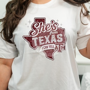May include: White t-shirt featuring a maroon glitter design in the shape of Texas. The design includes the words "She's from TEXAS" and "I CAN TELL A&M" in a decorative font. The shirt is a casual wear item.