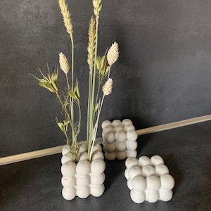 Könnte beinhalten: Drei graue Beton-Würfelvasen mit einem strukturierten Blasendesign. Die Vasen sind mit getrocknetem Weizen und anderen getrockneten Blumen gefüllt.