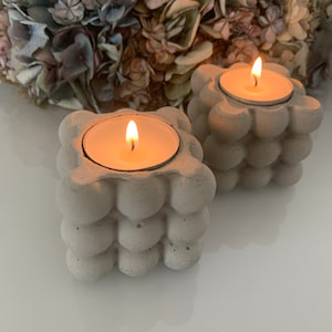 Op de afbeelding: Twee grijze betonnen kaarshouders met een getextureerd, bubbelvormig ontwerp. Elke houder heeft een kleine, ronde, witte kaars erin.