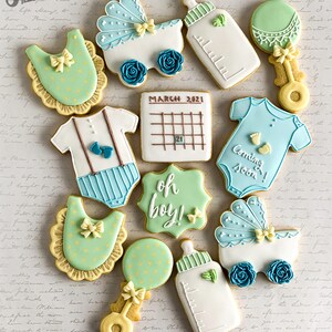 Puede incluir: Un conjunto de 12 galletas para baby shower decoradas con glaseado azul y verde. Las galletas tienen forma de babero, sonajero, biber&oacute;n, cochecito, mono, calendario y un cartel que dice "oh boy!"
