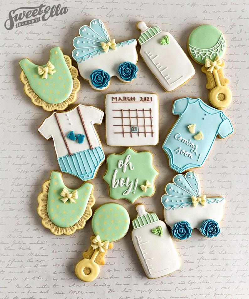 Puede incluir: Un conjunto de 12 galletas para baby shower decoradas con glaseado azul y verde. Las galletas tienen forma de babero, sonajero, biber&oacute;n, cochecito, mono, calendario y un cartel que dice "oh boy!"