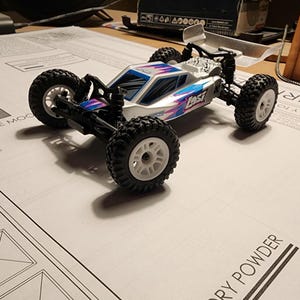 Peut inclure: Une voiture radiocommandée blanche et bleue avec des pneus noirs et un aileron en plastique transparent. La voiture porte l'inscription "Lost" sur le côté.