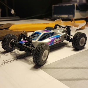 Peut inclure: Une voiture télécommandée argentée et bleue avec une aile en plastique transparente. La voiture a des pneus noirs et le mot "Losi" imprimé sur le côté.