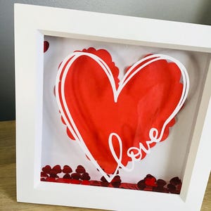 May include: A white framed shadow box featuring a red heart design with a white outline and the word "love". Red heart-shaped confetti is scattered at the bottom. The frame is a square shape.