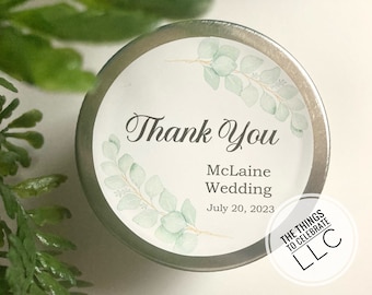 May include: A round silver container with a white label that says "Thank You" with a green watercolor eucalyptus design. The label also says "McLaine Wedding" and "July 20, 2023". The bottom of the label says "THE THINGS TO CELEBRATE LLC".