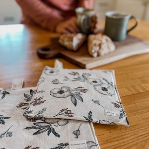 May include: Two white cotton pot holders with a floral pattern in shades of brown, black, and gray. The pot holders have a label that reads "The Northern Loom".