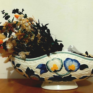 May include: A white ceramic bowl with blue and green floral designs. The bowl is filled with dried flowers and grasses, including orange and beige plumes.