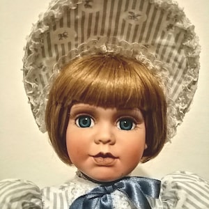 May include: A porcelain doll with blue eyes, brown hair, and a white and gray striped dress. The doll is wearing a white lace hat with a gray and white striped pattern.