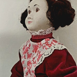 Puede incluir: Una muñeca de porcelana con el pelo castaño oscuro recogido en un moño, lleva un vestido de terciopelo rojo con ribete de encaje blanco y un delantal de estampado floral rosa.