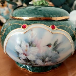 Puede incluir: Una caja de música de porcelana verde con un delicado diseño floral y un colibrí en vuelo. La caja está decorada con ribetes dorados y joyas rojas.