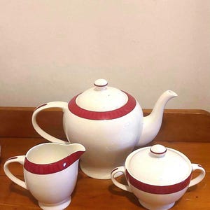 May include: A white ceramic tea set with a red band design. The set includes a teapot, creamer, and sugar bowl with a lid. The teapot has a curved spout and handle. The creamer and sugar bowl also have handles.