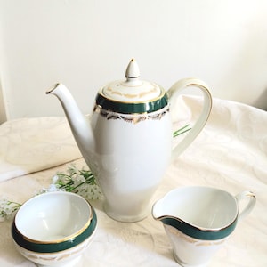 May include: A white porcelain coffee set consisting of a tall coffee pot with a lid, a sugar bowl, and a creamer. Each piece features a dark green band around the top rim, accented with a delicate gold leaf pattern and gold trim edges.