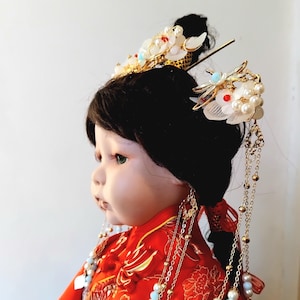 May include: A doll wearing a traditional Chinese red dress with gold embroidery. The doll has black hair styled with ornate gold and pearl hair accessories. Long gold chains with pearls and blue accents hang from the hairpiece.