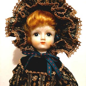 May include: A vintage porcelain doll with auburn hair and blue eyes, wearing a dark floral print dress and a matching bonnet with lace trim. The doll has a velvet collar and a teal ribbon.