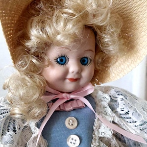 May include: A porcelain doll with blonde curly hair wearing a straw hat, a blue dress with white lace trim, and a pink ribbon around her neck.