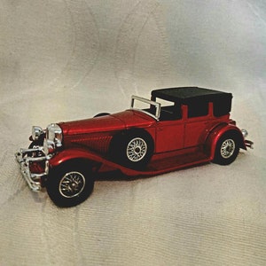 May include: A red toy car model of a 1930s era Duesenberg, a classic American luxury car. The car has a black roof and chrome accents. The model is detailed with a spare tire on the back and a detailed engine.