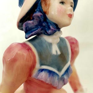 May include: A porcelain figurine of a woman in a pink dress and blue hat. The figurine has a detailed face with red hair and rosy cheeks. The dress has a blue bodice and the hat is trimmed with a blue ribbon.