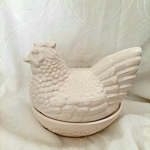 May include: A cream-colored ceramic chicken-shaped egg holder featuring a detailed molded texture on the feathers and a matching round base. The lid is shaped like a sitting hen with a comb on its head, resting securely on a bowl-shaped bottom.