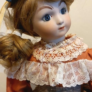 May include: A porcelain doll with blonde hair, blue eyes, and a painted smile. She is wearing a brown and white plaid dress with a lace collar and a brown hat.