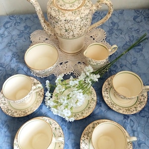 Può includere: Un servizio da tè composto da una teiera, una lattiera, una zuccheriera e quattro tazze con piattini abbinati. Ogni pezzo è color crema con un motivo floreale verde e oro e bordo dorato. Un rametto di fiori bianchi è posto al centro.