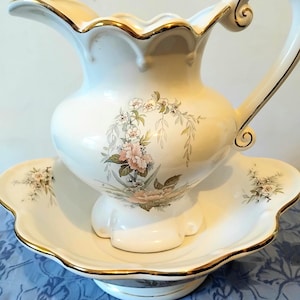 Könnte beinhalten: Ein Vintage-Set aus weißer Porzellankanne und Waschbecken. Das Set hat ein Blumendesign mit rosa und grünen Akzenten. Goldene Verzierungen säumen die Kanne und das Becken. Die Kanne hat einen geschwungenen Griff und einen Ausguss.