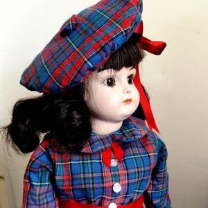 May include: A vintage porcelain doll wearing a blue, red, and green plaid dress, matching beret, and a red ribbon. The doll has black hair and is wearing a red belt. The doll's dress has white buttons.