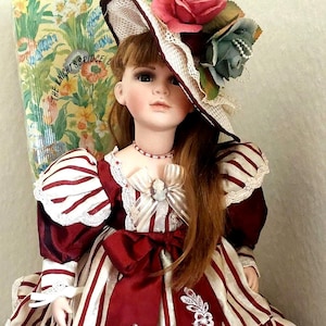 May include: A porcelain doll wearing a burgundy and cream striped dress with a matching hat adorned with flowers. The doll has long auburn hair and a delicate necklace. The doll is a collectible item.