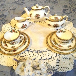May include: A white porcelain tea set featuring a floral pattern with red roses and green leaves, accented by gold trim. The set includes a teapot, a lidded sugar bowl, a creamer, two teacups, and two saucers, all arranged on a white crocheted doily.