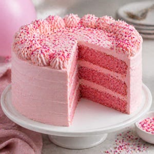 Può includere: Una torta rosa a più strati con una fetta rimossa, che rivela l'interno. La torta è glassata con crema al burro rosa e decorata con zuccherini e rosette rosa. Si trova su un alzatina bianca, con una piccola ciotola di zuccherini nelle vicinanze.