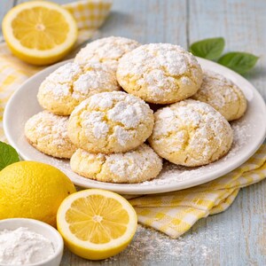 Può includere: Un piatto di biscotti al limone spolverati di zucchero a velo, insieme a limoni freschi e una piccola ciotola di zucchero a velo. I biscotti sono rotondi e dorati, con una superficie leggermente screpolata. La scena è ambientata su una superficie di legno azzurro chiaro con un tovagliolo a scacchi giallo.