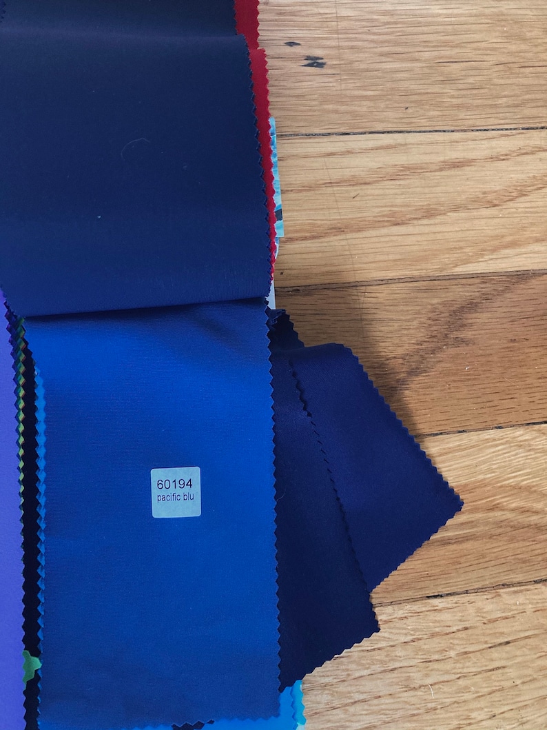 May include: A stack of fabric swatches in various shades of blue, including a dark navy blue, a medium blue, and a light blue. A small white label with the text "60194 pacific blu" is attached to the medium blue swatch.
