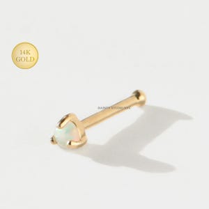 May include: A gold nose stud with a triangular opal stone. The stud is made of 14K gold and features a straight post. The opal has a pearlescent, iridescent quality. The brand name "DAINTY STUDIO NYC" is printed on the image.