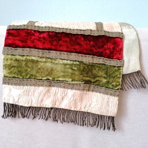 May include: A decorative textile with a cream-colored background, featuring horizontal bands of red and green velvet. The bands are separated by a gray trim and the bottom edge has a beaded fringe.