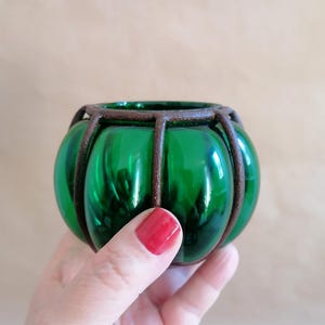 Könnte beinhalten: Ein grüner Glas-Kerzenhalter mit einem braunen Metallkäfig. Der Halter ist rund und hat eine glatte Oberfläche.