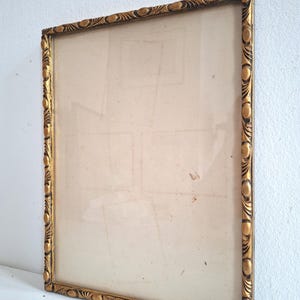 May include: A rectangular picture frame with a metallic gold finish and a decorative border featuring repeating oval and leaf-like patterns. The frame contains a clear glass pane and a light-colored backing board, presented in a vertical orientation.