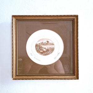 Pode incluir: Uma impressão quadrada emoldurada apresentando uma gravura circular em tons de sépia de uma paisagem rural com um degrau de madeira. A obra é intitulada The old stile e assinada pelo artista. A moldura é dourada com borda perolada.